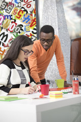 Wall Mural - Two diverse work colleagues smiling and writing down notes while sitting together at a table in a modern office