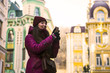 © Oleksandr - Pretty Brunette Girl Wearing Purple Winter Coat, Hat and Scarf, Walking by European Street at Winter, make Photos on Her Smartphone and Making Selfie