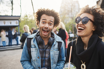 Wall Mural - Funny emotive young dark-skinned male with afro haircut making face at camera while spending time in park with best friend, drinking tea and enjoying lovely evening. Boyfriend makes girl laugh