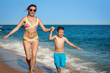 Mom and son are running along the sea shore. They are holding hands and moving towards the camera.