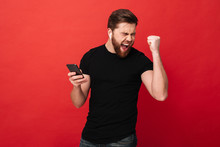 Delighted Bearded Guy Clenching Fist And Rejoicing With Closed Eyes While Using Mobile Phone And Wireless Earphones, Isolated Over Red Background