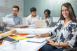 © Bojan - Smiling young business woman taking notes to clipboard while sitting in office with colleagues in the background.