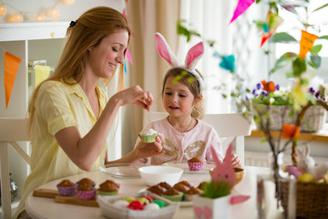 Mother and daughter celebrating Easter, cooking cupcakes, covering with glaze. Happy family holiday. Cute little girl in bunny ears.