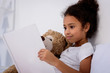 © LIGHTFIELD STUDIOS - adorable african american kid reading book and hugging teddy bear at home