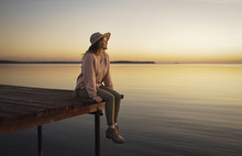 Woman Sitting On Dock Of Lake Admiring Sunset