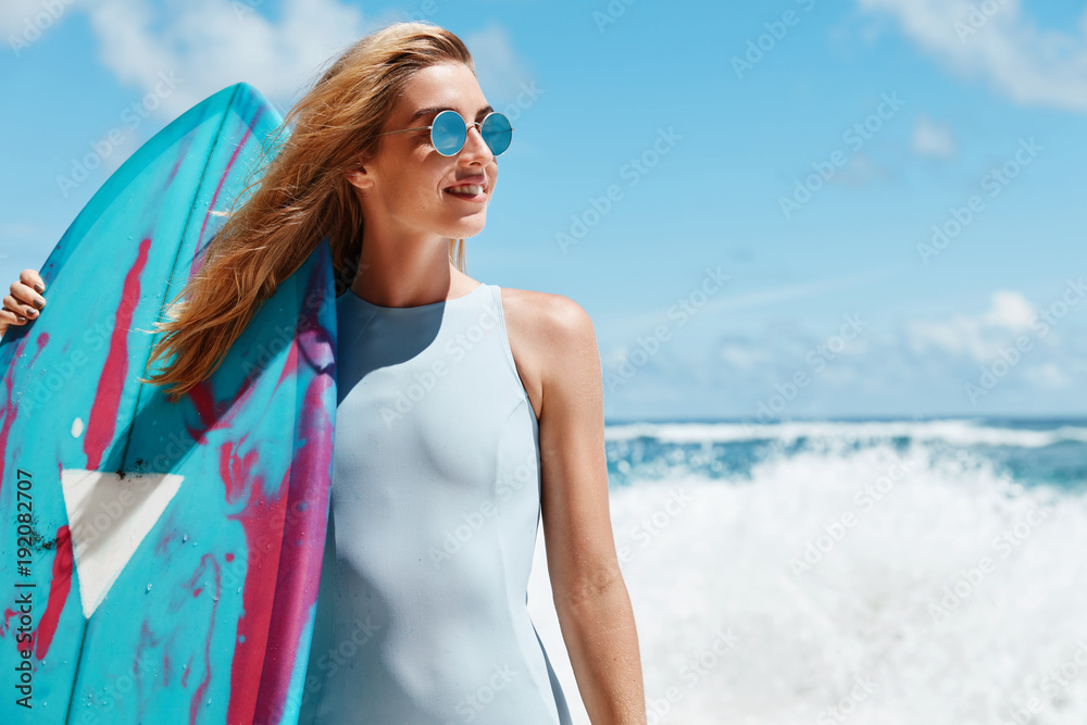Slim fit female model poses in swimwear, holds kite board, stands against stromy ocean, has appropriate weather conditions for surfing, daydreams of driving, has pleased satisfied expression - obrazy, fototapety, plakaty 