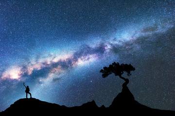 Milky Way, silhouette of woman and tree growing from the rock on the mountain at night. Space background with starry sky, beautiful galaxy and girl. Blue Milky Way, tree and woman. Universe. Adventure
