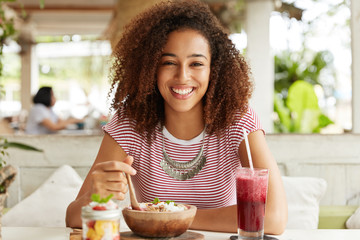 Adorabe dark skinned female with Afro hairstyle, wears casual striped t shirt, eats exotic sweet dish, drinks cocktail at outdoor cafeteria, smiles broadly, demonstrates white teeth, rests after work