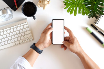 Wall Mural - Cropped shot top view of businessman hands using smartphone mockup at the white office desk. Blank screen mobile phone for graphic display montage