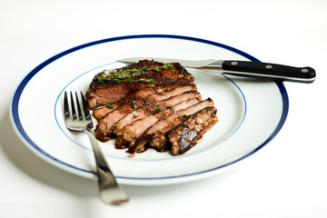 10oz Ribeye steak with thyme and smokey applewood seasoning Sliced and fanned on a blue rim plate on a white background With fork and steak knife