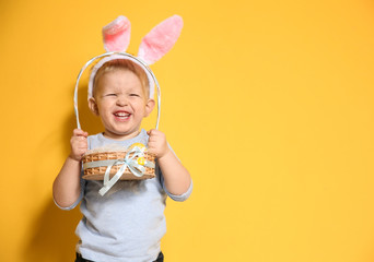 Cute little boy with bunny ears holding basket full of Easter eggs on color background