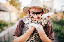 Happpy Young Bearded Farmer Holding Two Little Kitten In Hands Outdoor In Village With Abstract Background. Smiling Man In Glasses And Straw Hat Playing With Funny Cute Pets. Have Fun In Countryside.