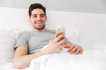 Sticker - Photo of happy awake man in t-shirt smiling while lying alone in bed with white linen, and holding golden cell phone