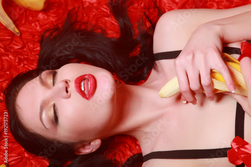 Zdjęcie XXL piękna seksualna brunetka dziewczyna ubrana w małą bieliznę, jedzenie bananów i leżąc na łóżku czerwony sam