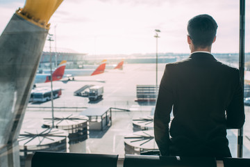 back of a young business man standing with the suitcase at the airport waiting for the flight .