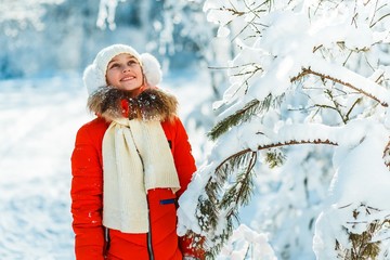 Beautiful teenage girl in a long red  down jacket white hat and a scarf having fun outside in a wood with snow in winter on a wonderful frosty sunny day.  active life consept