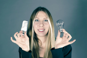 Wall Mural - Serious pretty business woman holding a light bulb on a grey background. Close-up face model with two kinds of light bulbs. Concept of saving money for electricity