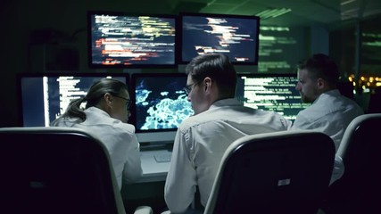 Rear view of three cyber security specialists talking at desk in front of multi-display workstation in dark IT office at night
