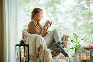 Young woman sitting home in a chair by the window with cup of hot coffee wearing knitted warm sweater. Cozy room decorated with lanterns and candles. Scenic view of pine trees in snow in window