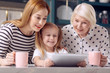 © zinkevych - Funnier together. Petite little girl showing a video on the tablet to her mother and grandmother while the women drinking coffee from pink cups