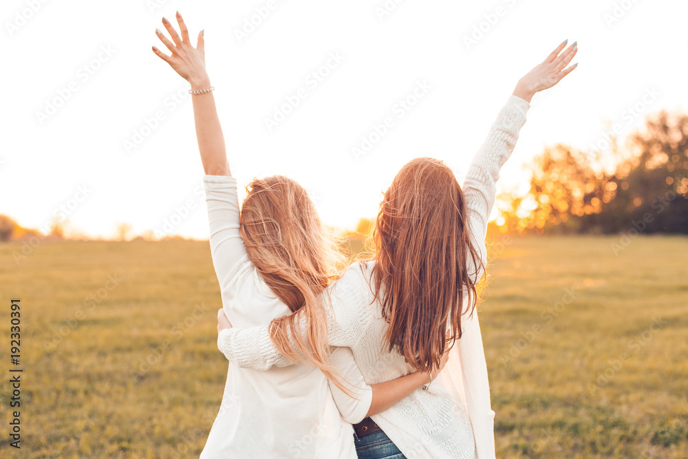 Two pretty girls raised their hands on a field at sunset. - obrazy, fototapety, plakaty 