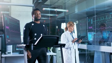 Wall Mural - male athlete walks on a treadmill with electrodes attached to his body while sport scientist supervi