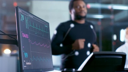 Wall Mural - male athlete runs on a treadmill with electrodes attached to his body while sport scientist holds ta