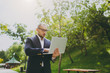 © ViDi Studio - Young successful smart businessman in white shirt, classic suit, glasses. Man standing and working on laptop pc computer in city park outdoors on nature background. Mobile Office, business concept.