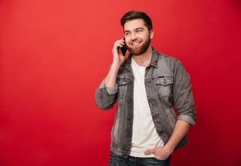 Canvas Print - Photo of handsome brunette man having call and speaking on smartphone with pleasure, isolated over red background