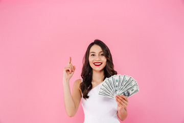 Poster - Gorgeous asian woman 20s holding fan of money in dollar cash and pointing finger upward on copyspace with smile, isolated over pink background