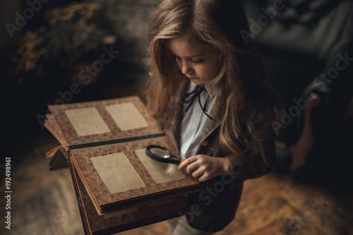 Fotografia Child girl in image of Sherlock Holmes stands in room and looks photoalbum with magnifier on background of old interior