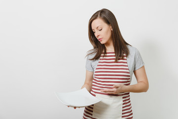 Young attractive smiling brunette caucasian housewife in striped apron, gray t-shirt isolated on white background. Beautiful housekeeper woman holding white empty plate. Copy space for advertisement.