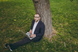 © ViDi Studio - Young successful businessman in white shirt, classic suit, glasses. Man sit on grass ground, work on laptop pc computer in city park on green lawn outdoors on nature. Mobile Office, business concept.