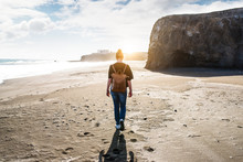 Back View Of Young Blonde Woman With Leather Backpack Walking On Empty Beach At Sunset