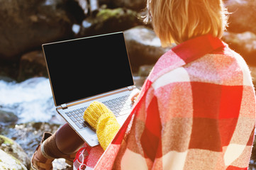 Mockup image of a girl wrapped in a plaid plaid with a laptop with a blank black desktop on her lap against the background of a mountain river and large stones. The concept of freelancing in freedom