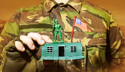 Sticker - Photo of a military man in camouflage jacket holding a soldier toy standing on barracks.