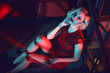 Beautiful glam blond woman with provocative make up wearing red short fitted sequin dress sitting on the stairs in the night club in colourful neon lights. Text space