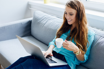 Attractive young woman sitting on sofa at home using laptop and holfing cup of coffee