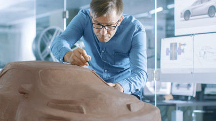 Wall Mural - Experience Automotive Designer with a Rake Sculpts Prototype Car Model from Plasticine Clay. He Works in a Modern Studio in a Major Automotive Company's Headquarters.