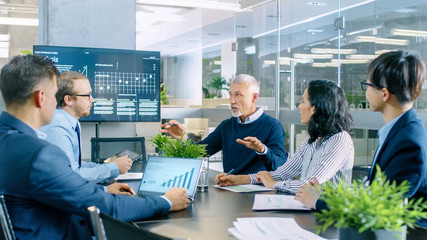 Senior Executive Explains Company's Vision and Potential to His Employees. They are Sitting at Big Table in Meeting Room. TV Screen on the Wall Shows Corporate Growth.