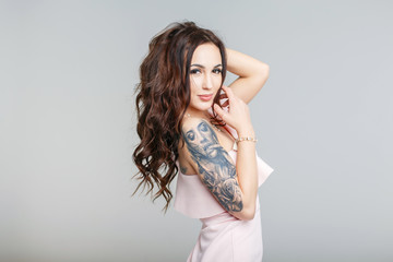 Beautiful young woman with stylish tattoo on hand in pink dress on gray background