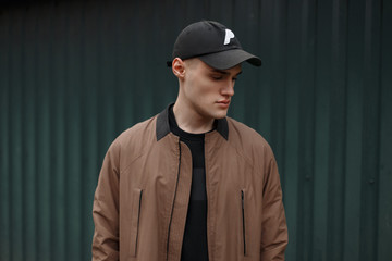 stylish fashion young man in a vintage coat and a fashionable cap stands near a green wall on the street