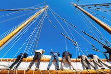 Crew Leans Over The Side Of A Big Sailboat To Raise The Heavy Sail On A Tall Ship At Sea. Takes Six Strong People To Hoist The Canvas Sail. Theme For Teamwork, Cooperation