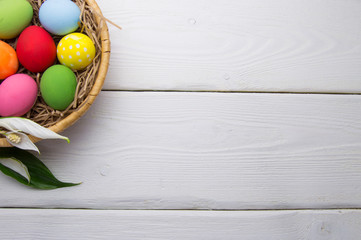 Wall Mural - Colored easter eggs on nest beasket with flower on white wooden background top white