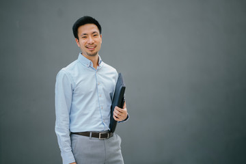 Portrait of happy smiling young businessman, standing on  a plain gray background while holding his leather folio. He is well-dressed in business casual and looks relaxed.