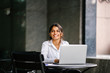 © Danon - An image of a young Asian Indian woman who types and works in a laptop and seeks a job. She wears a professional, crisp white shirt and she smiles during the day as she sits at a table.