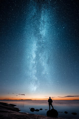 Wall Mural - Silhouette of a man standing alone on a rock by a sea and looking at sunset and stars of the Milky Way