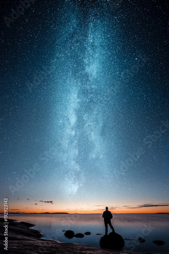 Fotomural Silhouette of a man standing alone on a rock by a sea and looking at sunset and