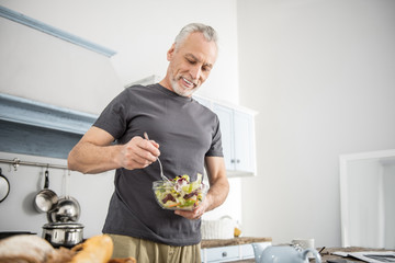 Wall Mural - Eat vitamins. Cheerful pensioner being in the kitchen and keeping smile on his face while mixing salad