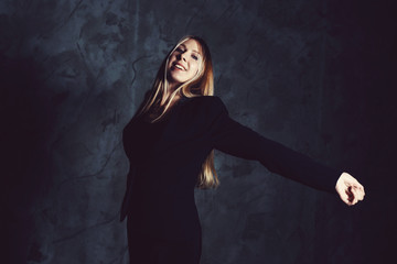 Attractive pretty young model posing against a dark studio background with copy space looking. Atomic blonde woman stands with widely spread arms. Cold cinematic style